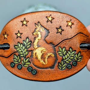 K&ouml;nnte beinhalten: Eine braune Haarklammer aus Leder mit einem goldfarbenen Design aus Fuchs, Sternen und Bl&auml;ttern. Die Klammer hat eine schwarze Metallstange zum Festhalten der Haare.