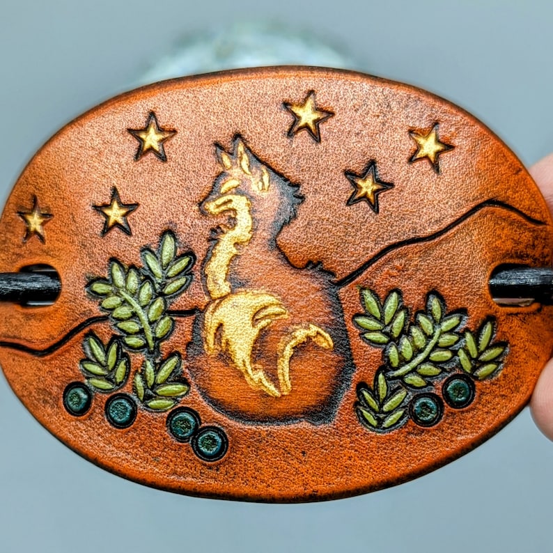 K&ouml;nnte beinhalten: Eine braune Haarklammer aus Leder mit einem goldfarbenen Design aus Fuchs, Sternen und Bl&auml;ttern. Die Klammer hat eine schwarze Metallstange zum Festhalten der Haare.