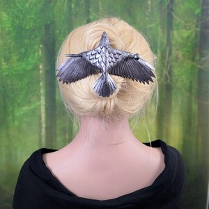 May include: A silver-colored hair clip in the shape of a bird with outstretched wings. The bird's wings and tail feathers are detailed with textured lines. The hair accessory is set against a backdrop of a person's blonde hair and a green forest.