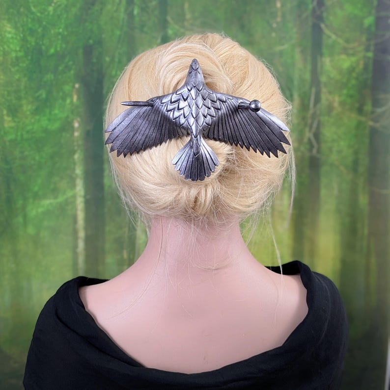May include: A silver-colored hair clip in the shape of a bird with outstretched wings. The bird's wings and tail feathers are detailed with textured lines. The hair accessory is set against a backdrop of a person's blonde hair and a green forest.