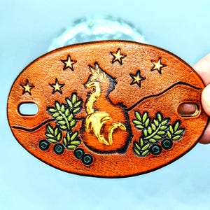 K&ouml;nnte beinhalten: Ein brauner Leder-Schl&uuml;sselanh&auml;nger mit einem goldenen Fuchs-Design, gr&uuml;nen Bl&auml;ttern und blauen Beeren. Der Fuchs ist von Sternen umgeben.