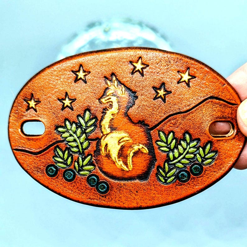 K&ouml;nnte beinhalten: Ein brauner Leder-Schl&uuml;sselanh&auml;nger mit einem goldenen Fuchs-Design, gr&uuml;nen Bl&auml;ttern und blauen Beeren. Der Fuchs ist von Sternen umgeben.
