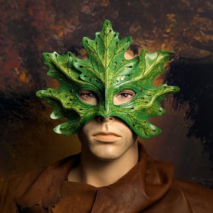 May include: A green leaf-shaped mask featuring layered foliage detailing with intricate vein patterns and small circular cutouts. The mask is worn over the eyes and upper face, displaying a textured surface with shades of light and dark green.