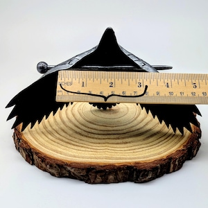 May include: A black metal decorative bird sculpture with a wingspan of approximately 4 inches, resting on a natural wood slice. The bird has a detailed, textured design and is set against a white background.