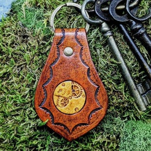 May include: A brown leather keychain with a gold-colored gear design. The keychain is attached to a silver key ring.