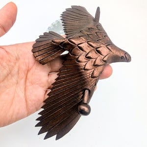 May include: Bronze-colored eagle sculpture in mid-flight, with detailed feather and wing designs. The sculpture is held in a hand, showcasing its size and intricate craftsmanship. The eagle's beak is slightly open, and the wings are spread wide.