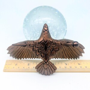 May include: Bronze-colored bird sculpture with outstretched wings, detailed feather design, and a small ball at each wingtip. The sculpture is placed on a ruler, measuring approximately 6 inches in length. A clear glass sphere is in the background.
