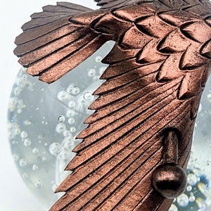 May include: Close-up of a bronze-colored bird figurine with detailed wing feathers. The bird is positioned in front of a clear glass globe filled with bubbles, creating a whimsical effect. The figurine's texture and color are prominent.