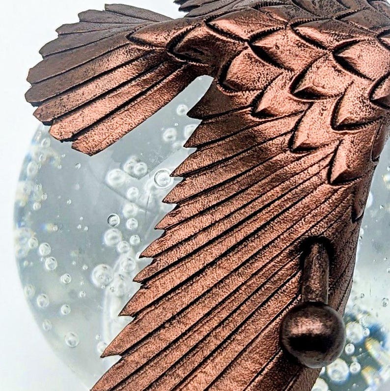 May include: Close-up of a bronze-colored bird figurine with detailed wing feathers. The bird is positioned in front of a clear glass globe filled with bubbles, creating a whimsical effect. The figurine's texture and color are prominent.