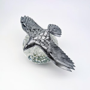 May include: A silver-colored, detailed sculpture of a bird in flight, perched atop a clear glass orb with bubble inclusions. The bird's wings are outstretched, showcasing intricate feather detailing. The sculpture is set against a white background.