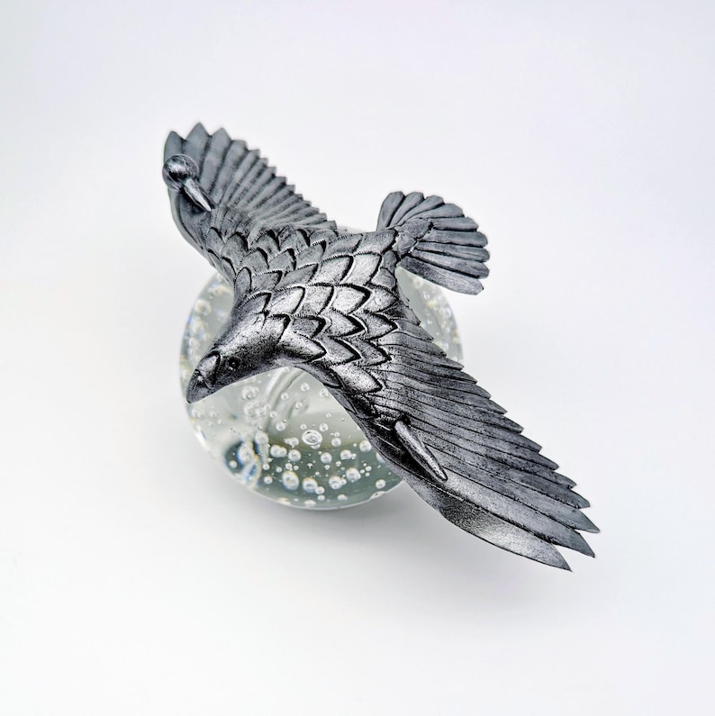 May include: A silver-colored, detailed sculpture of a bird in flight, perched atop a clear glass orb with bubble inclusions. The bird's wings are outstretched, showcasing intricate feather detailing. The sculpture is set against a white background.