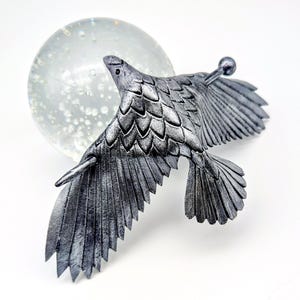 May include: A silver-colored metal bird sculpture in flight, with detailed feather textures. The bird is positioned in front of a clear glass orb, creating a striking contrast. The sculpture has a small, round detail extending from its back.