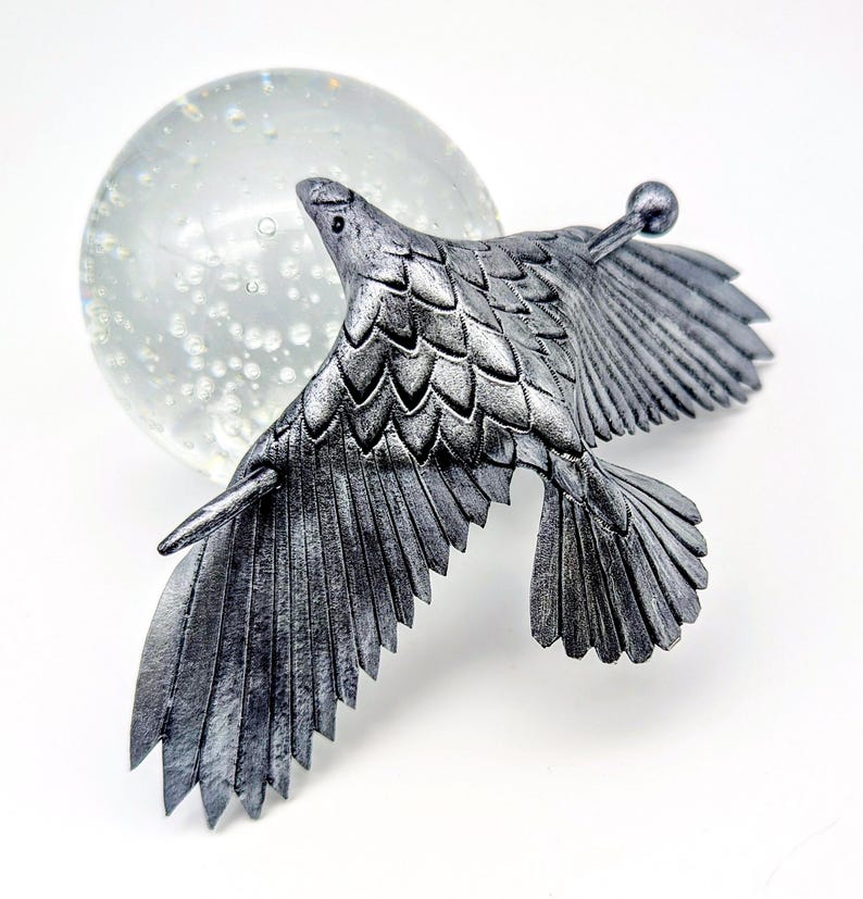 May include: A silver-colored metal bird sculpture in flight, with detailed feather textures. The bird is positioned in front of a clear glass orb, creating a striking contrast. The sculpture has a small, round detail extending from its back.