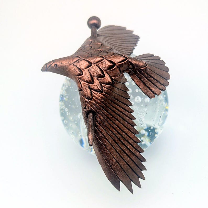 May include: A bronze-colored metal eagle sculpture in mid-flight. The eagle has detailed feather patterns and is mounted above a clear glass sphere with blue and white accents. The sculpture has a small ball at the top.
