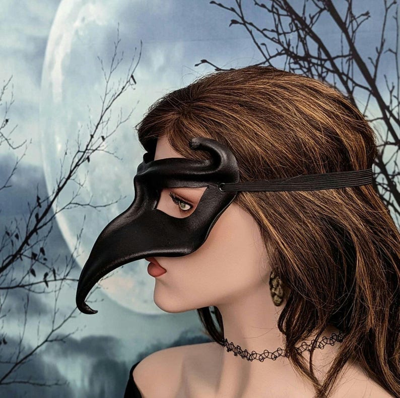 May include: A black leather masquerade mask with a long, curved beak. The mask has a single eye hole and is worn by a person with long brown hair.