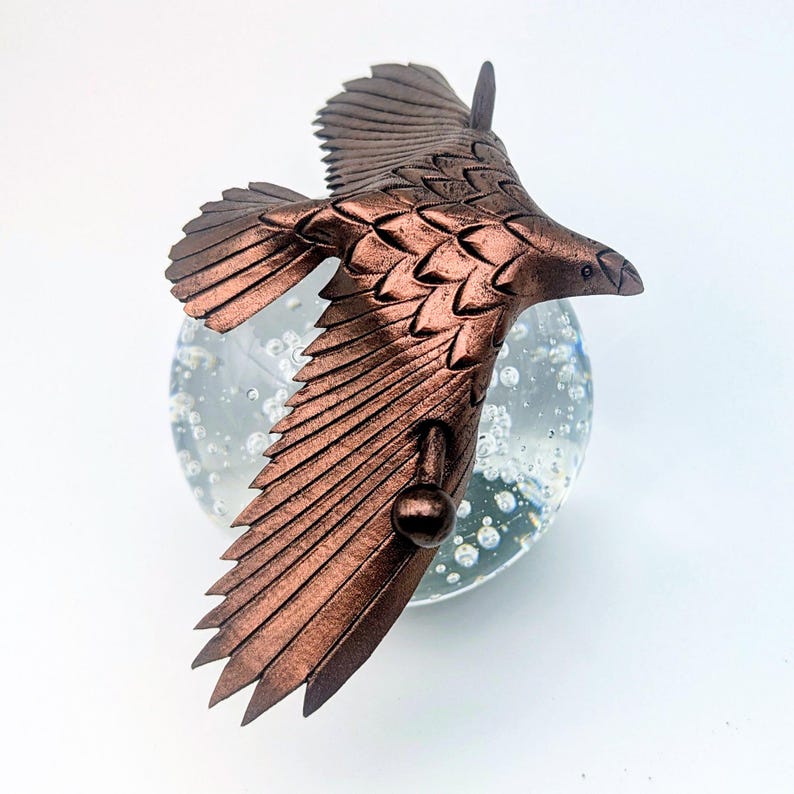May include: A bronze-colored eagle sculpture in mid-flight, with detailed feathering. The eagle is positioned above a clear glass orb with bubbles, creating a dynamic and artistic composition. The sculpture is likely a decorative item.