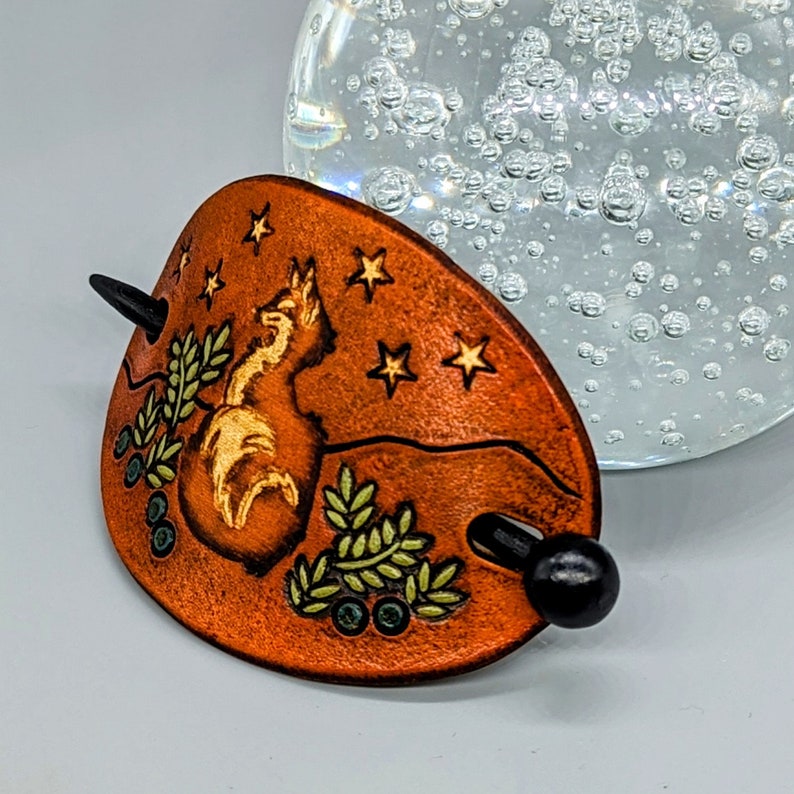K&ouml;nnte beinhalten: Eine braune Haarklammer aus Leder mit einem Fuchsdesign und Sternen. Die Klammer hat einen schwarzen Holzstab, der durch das Leder geht.
