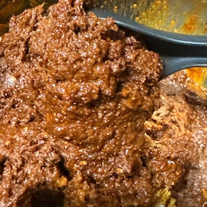 May include: Close-up of a dark brown, textured food mixture in a bowl. A black spatula is partially visible, stirring the thick, chunky substance. The food appears to be a sauce or paste, with a rich, glossy appearance.