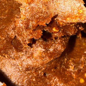 May include: Close-up of a textured, dark brown food item with a slightly grainy appearance. The surface has a rough, uneven texture with visible layers and small, light-coloured particles. The food appears to be moist and possibly spreadable.