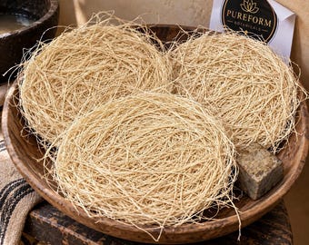 May include: Three circular, light tan, fibrous nesting pads made of natural plant fibers arranged in a large, dark brown, round wooden bowl. A small, rectangular, mottled gray bar of soap sits in the bowl next to a white card labeled PUREFORM BOTANICALS.