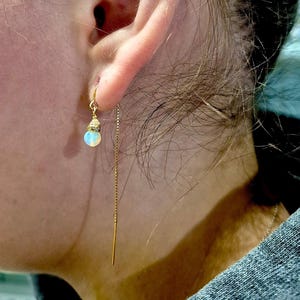 May include: A close-up of a gold earring with a small, round, opalescent bead and a delicate chain that dangles down. The earring is attached to a gold hook. The earring is worn in the ear.