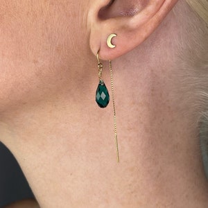 May include: Gold dangle earrings with a dark green teardrop-shaped crystal and a thin gold chain. The earrings are shown on a person's ear.
