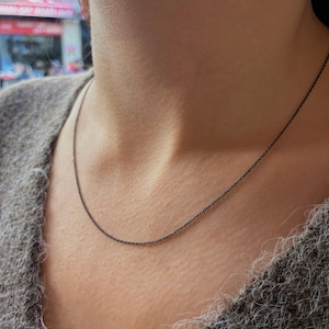 May include: A thin, dark-colored metal chain necklace with small, uniform oval links. The necklace rests against the neck, featuring a simple, minimalist design with a smooth, matte finish that provides a subtle contrast against the skin.