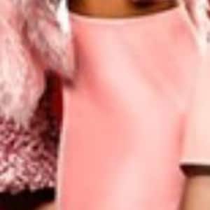 May include: A woman in a pink outfit, including a crop top and skirt, is surprised. She is wearing a pink fur jacket and high heels. Another person is holding a knife. The background is red.