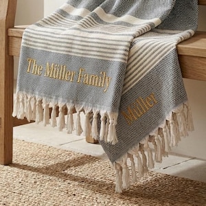 Op de afbeelding: Een grijs-witte gestreepte deken met de geborduurde tekst "The Müller Family" en franjes. De deken is over een houten bank gedrapeerd. Bruine sandalen liggen op een geweven vloerkleed.