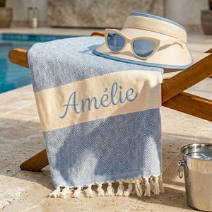May include: A blue and white patterned beach towel embroidered with the name "Amélie", a cream-coloured visor with blue trim, and sunglasses are on a wooden chair by a pool. A small metal bucket is also present.