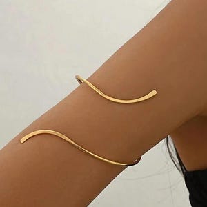 May include: A gold-toned arm cuff featuring a smooth, continuous wave-like design that wraps around the upper arm. The metal band has a polished finish and a curved, serpentine shape with tapered ends, creating an open-ended, flexible structure.