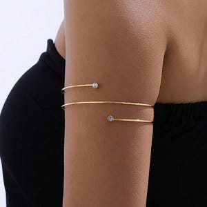 May include: Gold-coloured arm cuff with two parallel bands. Each band has a small, clear, round gemstone. The jewellery is worn on the upper arm. The background is a dark colour.