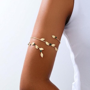 May include: Gold-coloured arm cuff with a delicate leaf design and small green accents. The jewellery is worn on the upper arm and has a minimalist, nature-inspired aesthetic. The cuff is a stylish accessory.