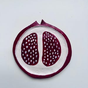 May include: A circular, hand-painted decorative dish shaped like a halved pomegranate. The outer rim is a deep burgundy red, while the centre is white with two symmetrical, dark red sections filled with small, white, teardrop-shaped seed details.