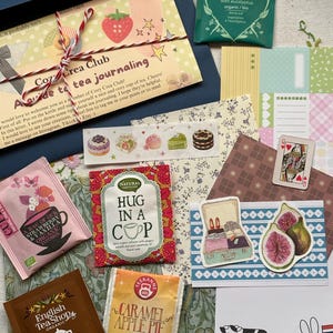 Może przedstawiać: Zestaw upominkowy z artykułami papierniczymi i herbatą, zawierający przewodnik Cozy Crea Club, herbatę Pukka Breathe In, herbatę English Tea Shop Rooibos Cocoa & Vanilla, herbatę Teekanne Caramel Apple Pie, herbatę Natural Temptation, taśmę washi, naklejki i papier.