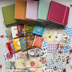 Könnte beinhalten: Fünf Notizbücher mit festem Einband in Oliv, Senf, Lavendel, Minze und Magenta mit Gummibändern, umgeben von verschiedenen Teebeuteln, dekorativen Aufklebern, Alphabetbögen, Washi-Tape-Rollen und einem Leitfaden mit dem Titel A guide to tea journaling.