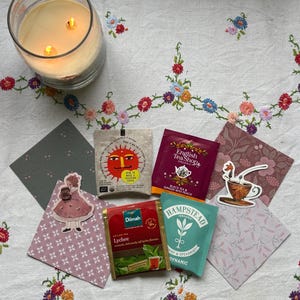 May include: A glass candle with two lit wicks is next to tea bags and decorative cards on a floral embroidered tablecloth. Tea bag brands include Dilmah Lychee, English Tea Shop, and Hampstead Organic Mint & Spearmint.