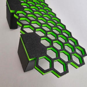 May include: A 3D printed object featuring a black hexagonal grid pattern with a vibrant neon green interior layer. The structure includes two solid black rectangular support blocks attached to the underside of the honeycomb mesh surface.