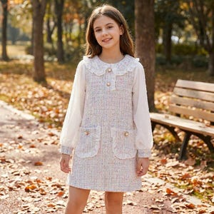 May include: A young person wearing a white and pastel-colored tweed dress with a Peter Pan collar and long, sheer sleeves. The dress features two front pockets with ruffled edges and gold-colored buttons. The background shows a park setting with trees and a bench.