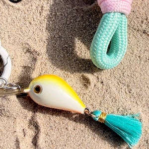 Könnte beinhalten: Ein Schlüsselanhänger-Accessoire mit einem weißen und gelben tropfenförmigen Anhänger mit einem metallischen Auge, befestigt an einer kleinen türkisfarbenen Quaste mit Goldfaden. Der Anhänger ist mit einer hellblauen Webseilschlaufe und einem rosa geflochtenen Seilteil verbunden.