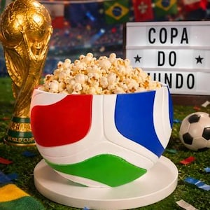 May include: A white bowl shaped like a soccer ball with red, blue, and green panels filled with popcorn. Beside it sits a gold-colored trophy replica and a light box displaying the text COPA DO MUNDO, all set against a blurred stadium background.