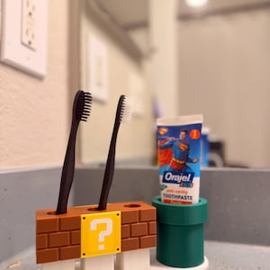May include: A toothbrush holder featuring a brown brick design with a yellow question mark block and a green pipe section. Two black toothbrushes are inserted into the brick base, and a tube of Orajel kids anti-cavity toothpaste sits in the pipe.