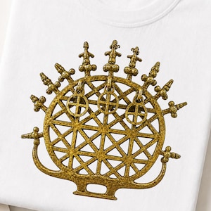 May include: A white t-shirt featuring a gold-colored graphic of a circular, lattice-patterned sun disc. The design includes three dangling circular charms with cross symbols and several small, stylized figures extending from the top edge.