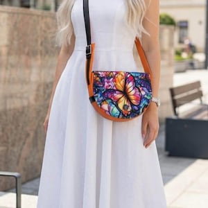May include: A colorful, half-moon shaped crossbody bag with a butterfly design. The bag features a vibrant butterfly illustration in shades of orange, blue, and purple. The bag has a brown leather strap and trim, and a black strap.