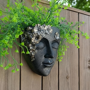 Puede incluir: Una maceta de pared gris oscuro con forma de rostro humano y ojos cerrados. La maceta presenta un grupo de flores de tono dorado metálico que caen por un lado. Un follaje artificial de color verde brillante sobresale por la parte superior.