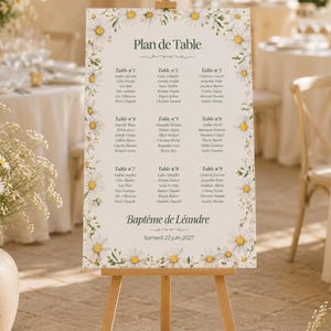 May include: A rectangular wedding seating chart sign on a wooden easel, featuring a white background with a border of white and yellow daisy flowers. The text reads Plan de Table, Baptême de Léandre, and Samedi 22 juin 2027, with nine tables listed.