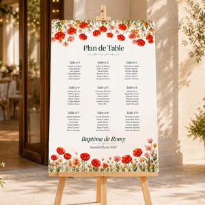 May include: A white wedding seating chart sign on a wooden easel, featuring a floral border of red poppies and small wildflowers at the top and bottom. The text reads Plan de Table, Baptême de Romy, and Samedi 22 juin 2027, with nine tables listed.