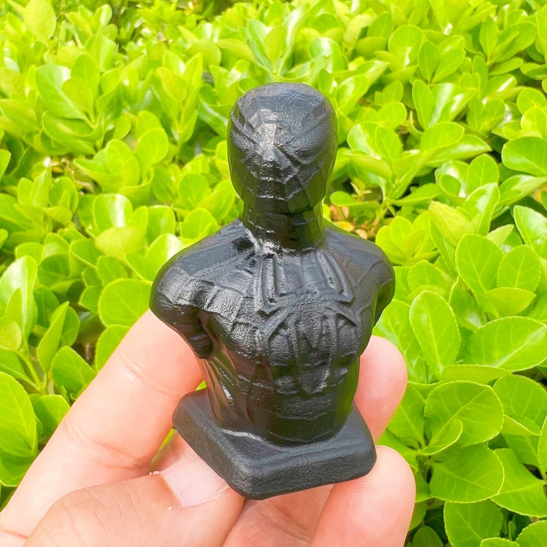May include: A black bust of a superhero with a spider-web design on the head and chest. The bust is on a small rectangular base. The background is a vibrant green bush.