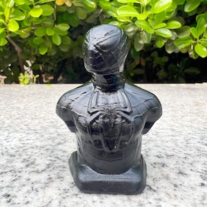 May include: A black bust of a superhero, featuring a detailed spider emblem on the back. The sculpture is set against a backdrop of green foliage and a textured surface. The bust is a solid, dark color.
