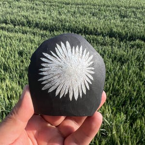 May include: A black, hand-carved stone with a white floral design. The flower has multiple petals radiating from a central point. The stone is held in a hand, with a green field in the background.